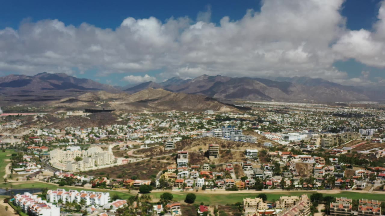 A Los Cabos aerial frame shows clustered white buildings and homes across a valley, with green fairway-like areas, brown hills, mountain ridges, broken cloud cover, and no visible people.