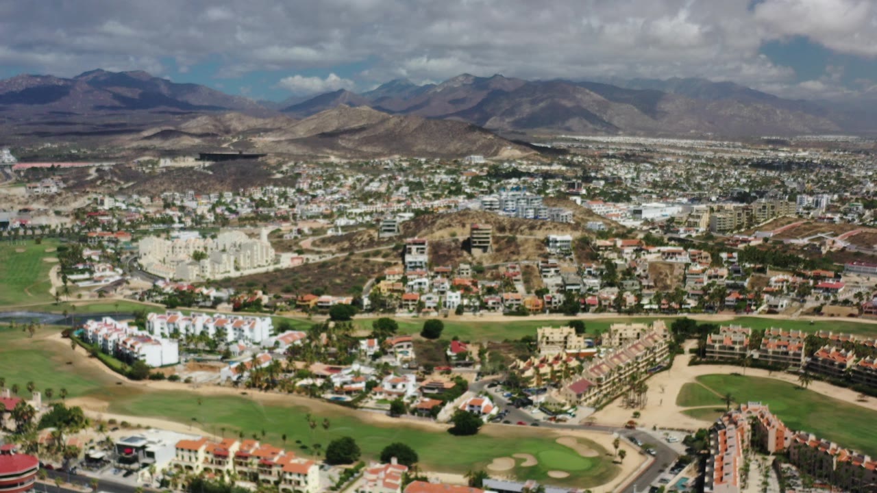 Wide aerial view of Los Cabos neighborhoods, golf greens, roads, hills, and clouds.