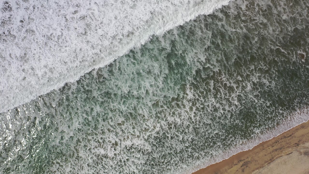 Overhead view of white surf spreading across green water toward tan sand in Los Cabos, Mexico, with no visible people.