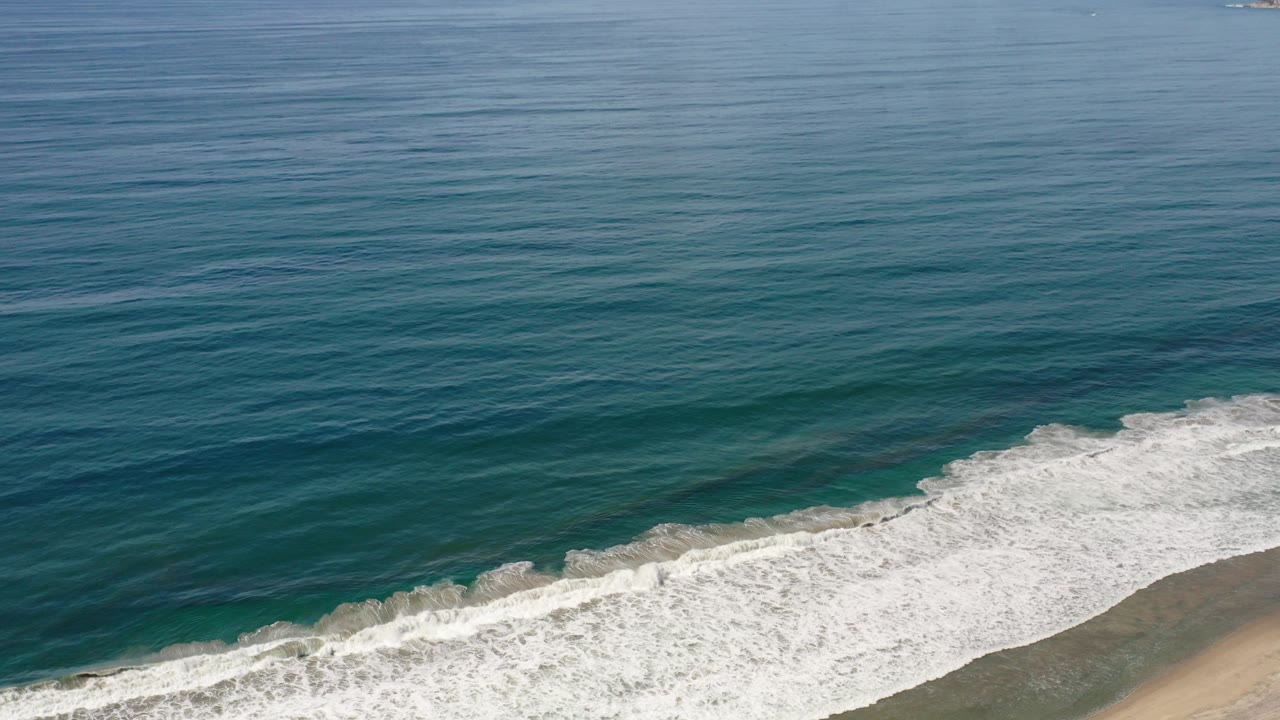 A wide elevated view in Los Cabos, Mexico shows blue-green ocean water and white surf reaching a sandy shore, with no visible people.