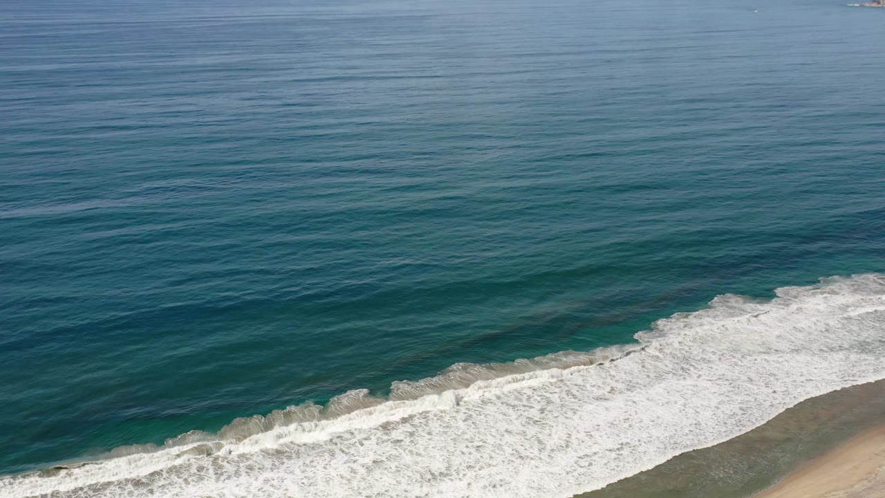 The surf band stretches diagonally across the lower frame in Los Cabos, Mexico, with darker blue water beyond it and no visible people.