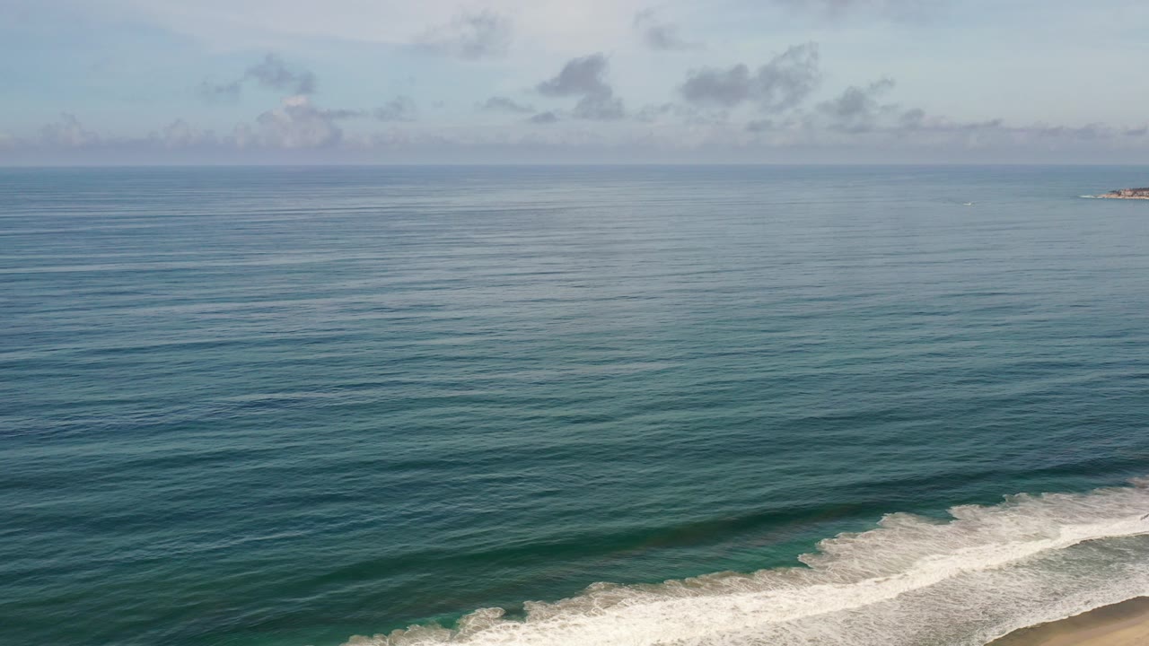 Opening frame of Los Cabos, Mexico coastline with blue-green ocean, white surf at the lower edge, pale clouds above the horizon, and no visible people.