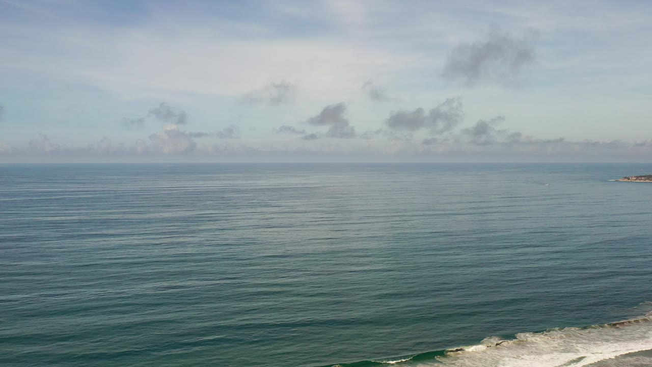 Wide Los Cabos ocean view with pale clouds, blue-green water, a distant rocky landform near the horizon, white surf at the lower edge, and no visible people.