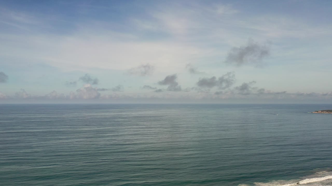Los Cabos ocean horizon under a hazy blue-gray sky, with calm water, small distant offshore rocks near center right, faint surf at the lower edge, and no visible people.