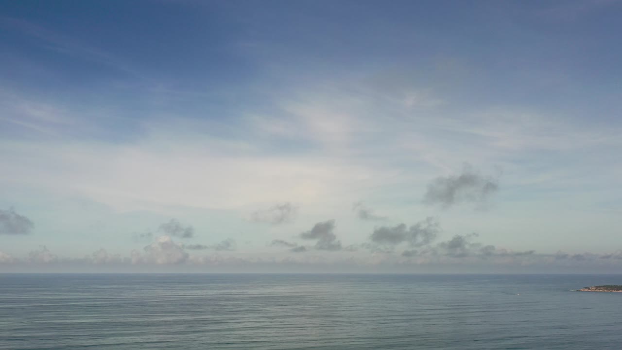 Wide Los Cabos ocean view at the start of the clip, with blue-gray cloud layers, a calm horizon, a small rocky islet near the right side, and no visible people.