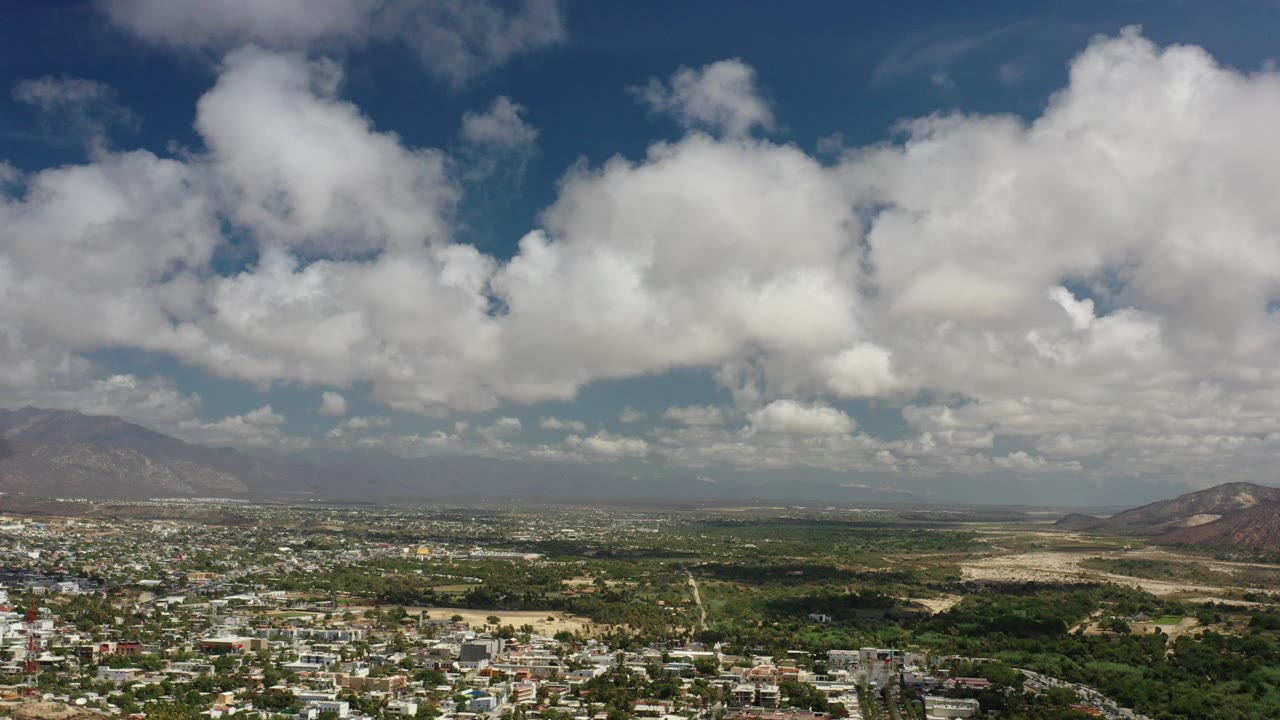 Opening view over Los Cabos, Mexico with broad white clouds, a hazy mountain ridge, scattered buildings, green scrubland, and no visible people.
