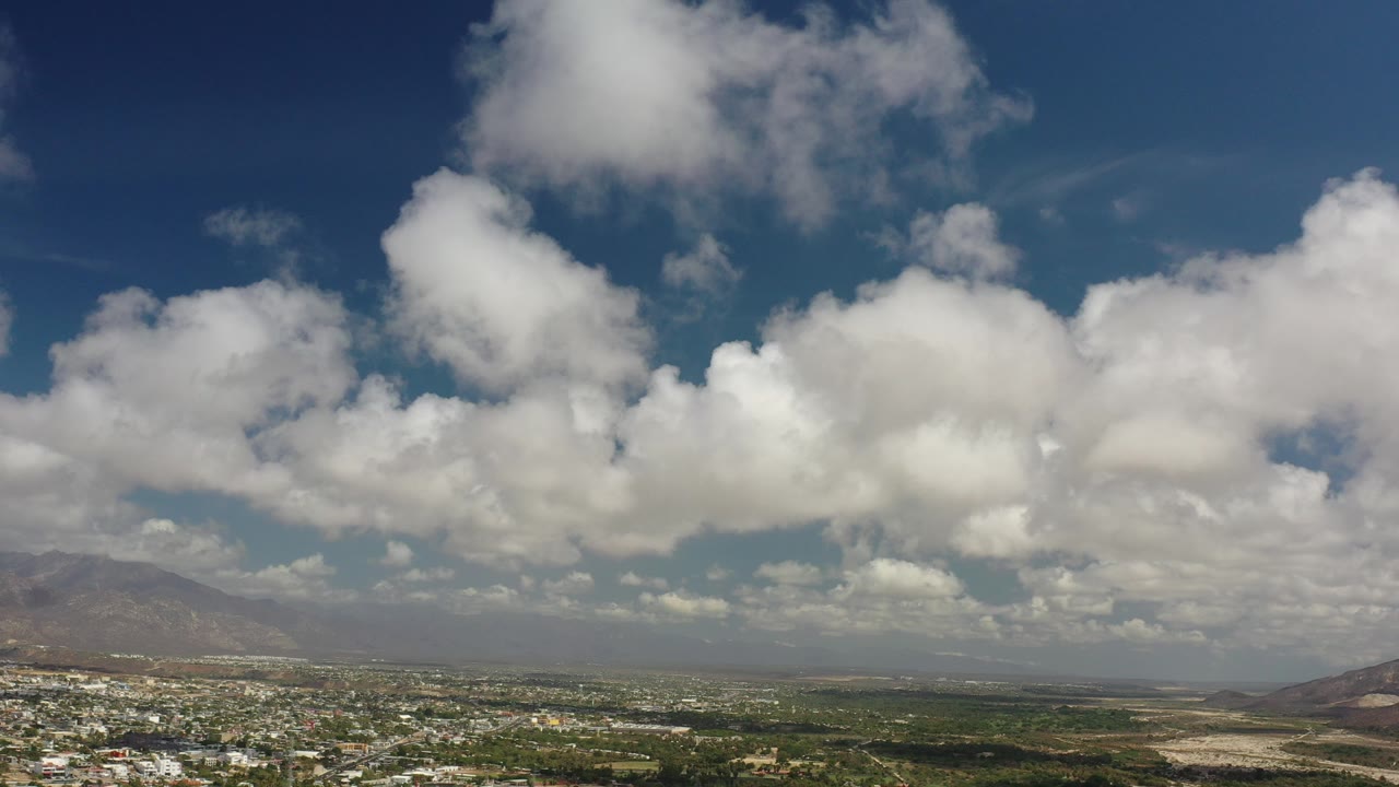 At 0:00, a wide Los Cabos, Mexico view shows large white clouds across a blue sky above hazy hills, scrubland, and low buildings, with no visible people.