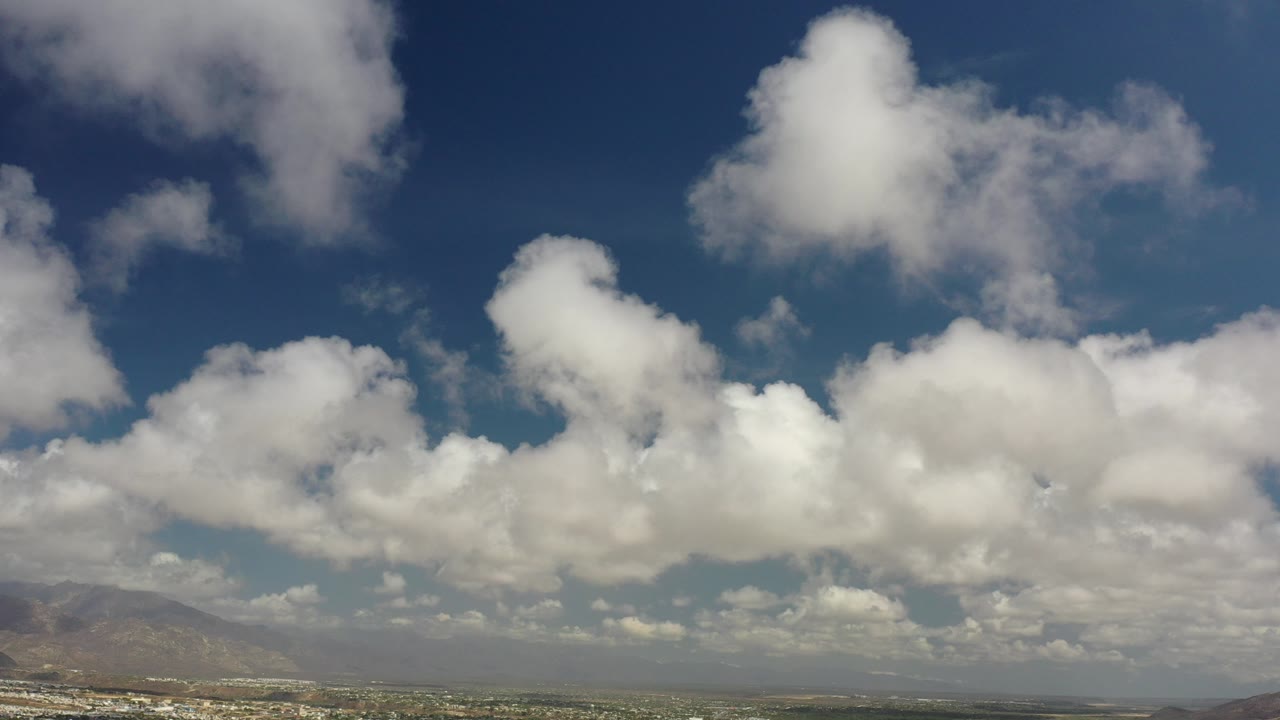 Opening frame of white clouds spread across a blue Los Cabos sky above a thin band of distant mountains and coastline, with no visible people.