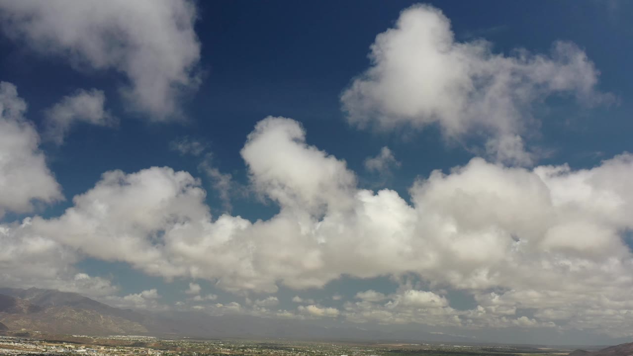 White cumulus clouds sit high over hazy mountain peaks and a narrow coastal horizon in Los Cabos, with no visible people.