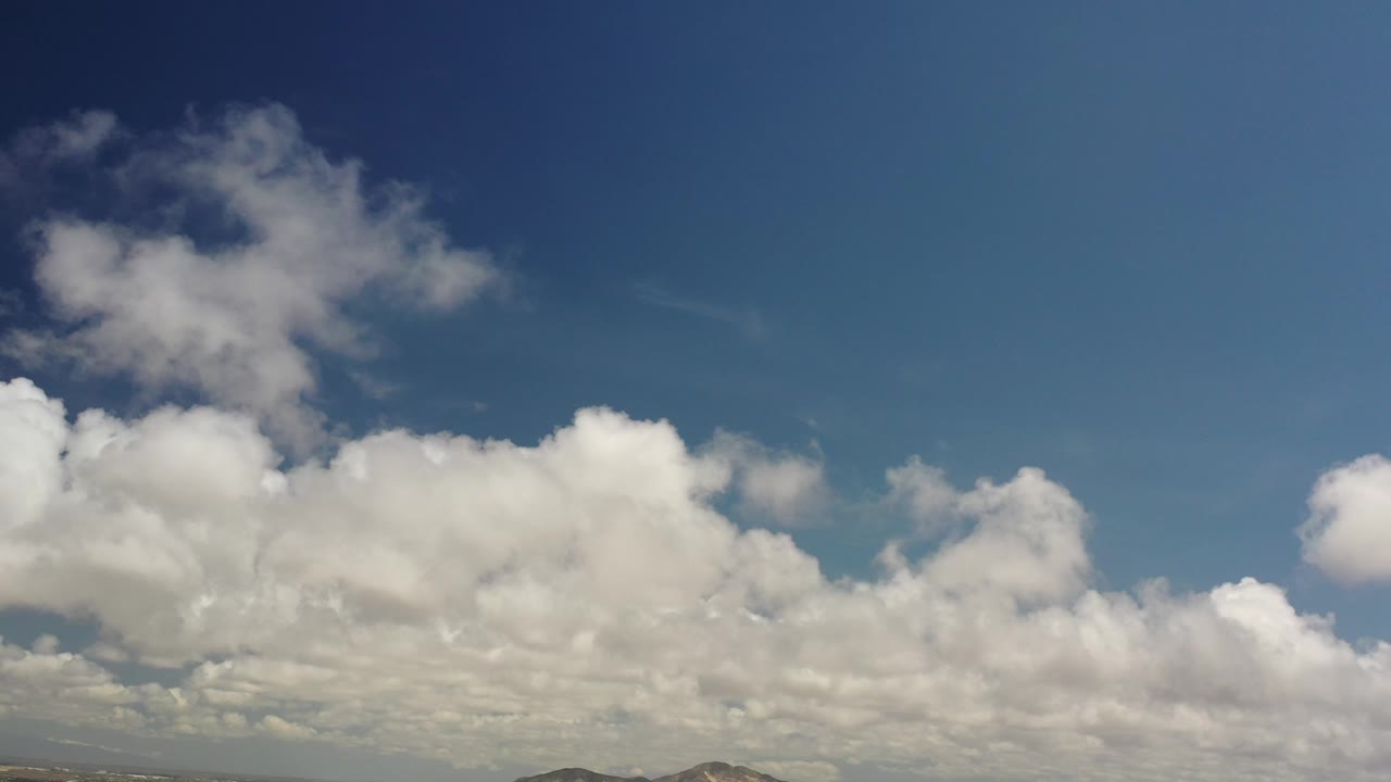 White clouds stretch across a blue sky over low hills.