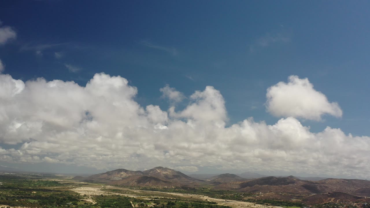 Opening frame of Los Cabos, Mexico with a blue sky, dense white clouds, brown hills, green lowlands, and no visible people.