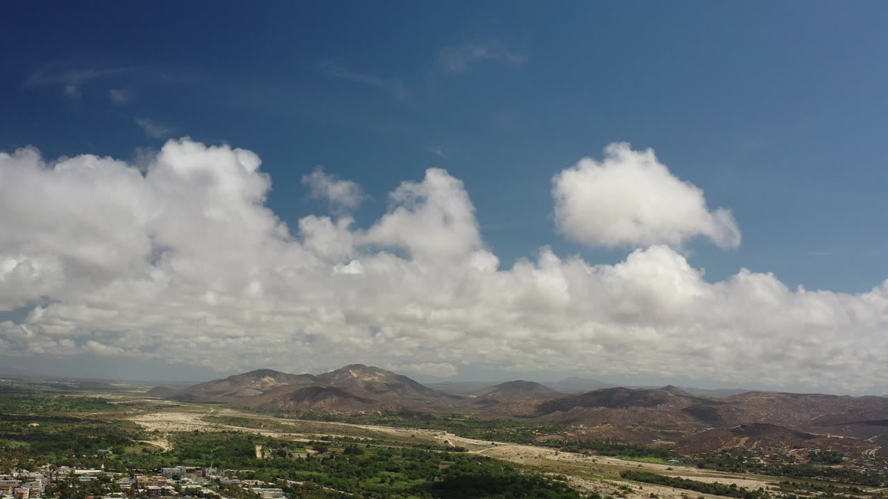 Opening wide view in Los Cabos, Mexico, with large white clouds above low mountains, green scrub, pale ground, and no visible people.