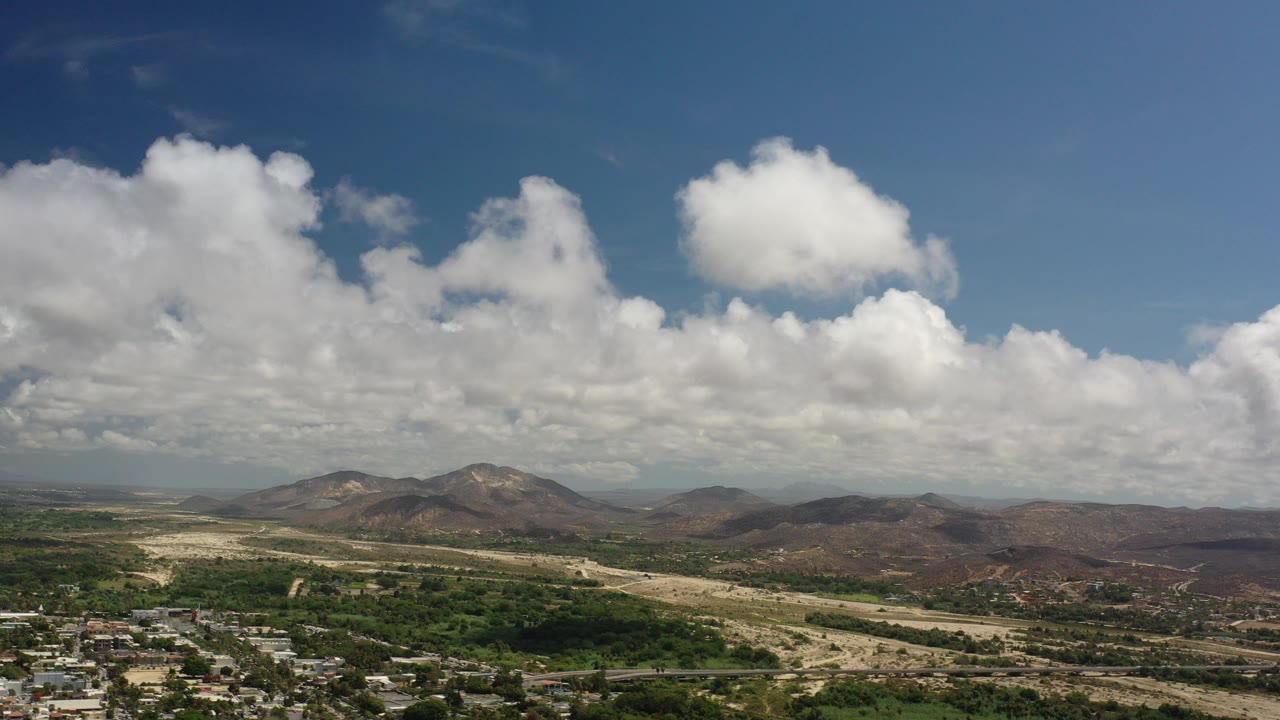 Wide aerial view of hills, valley, town edge, and cloud-filled sky.