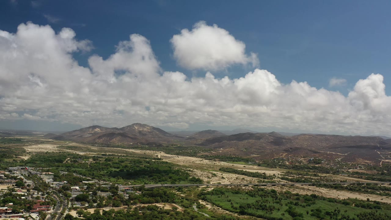 Los Cabos aerial frame with a road and scattered buildings lower left, green fields across the foreground, distant brown ridges, a blue sky, and no visible people.