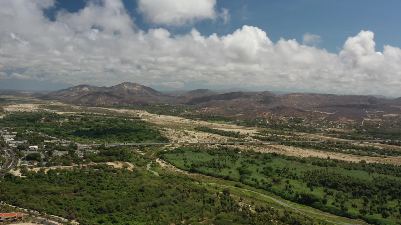 Opening aerial frame over Los Cabos, Mexico, with green scrub and fields below, pale dry terrain in the middle distance, low mountains on the horizon, and no visible people.