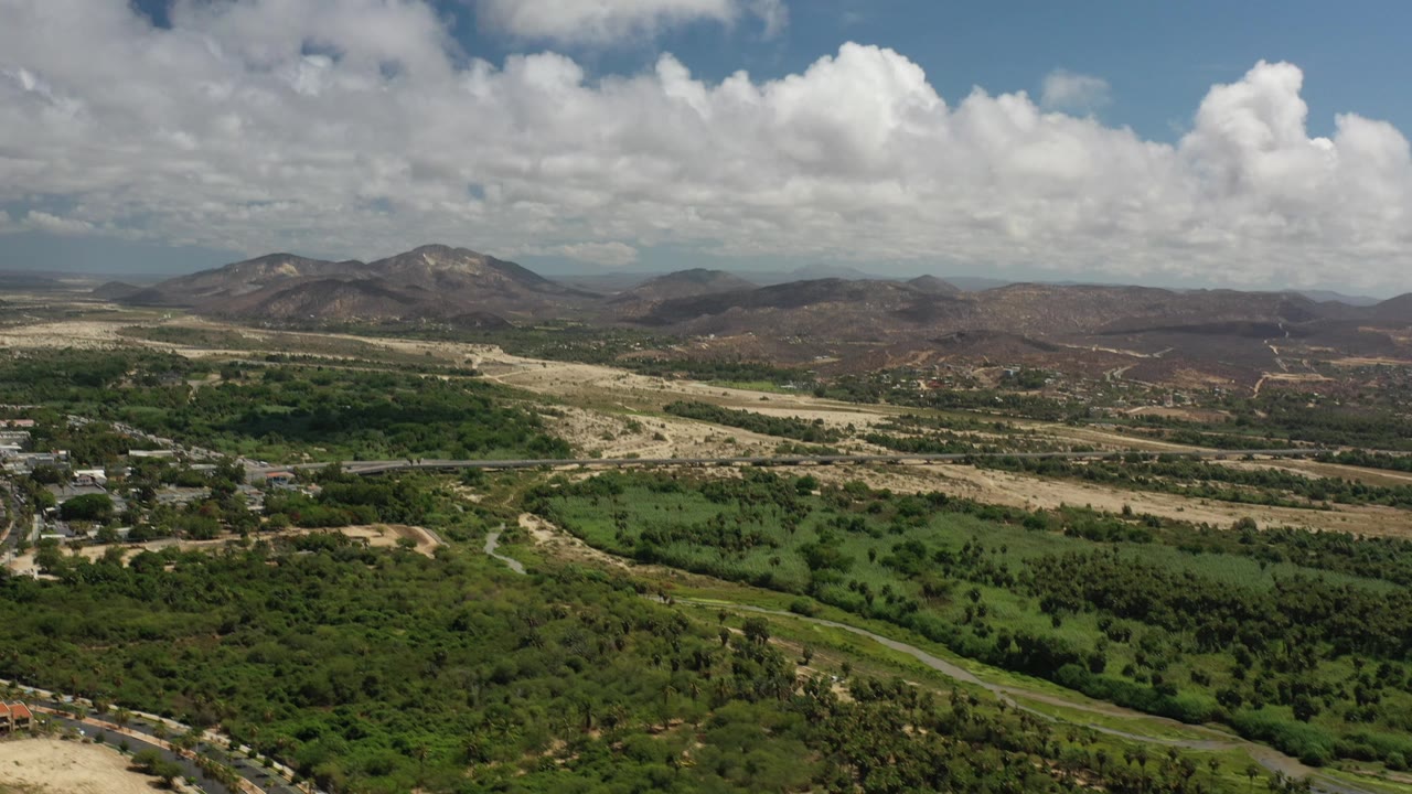 Elevated view across green fields, a road network, dry terrain, mountains, and clouds.