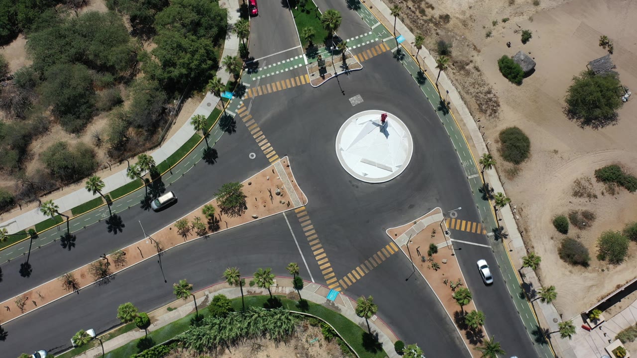 Overhead Los Cabos, Mexico frame with the roundabout near center, one white car along the right road, a dark car at lower left, sidewalks, palms, and sandy open ground with no visible people.