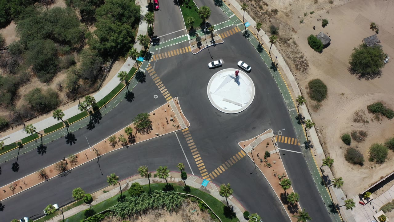 Overhead frame in Los Cabos, Mexico showing two white cars approaching a circular roundabout with marked crosswalks, sidewalks, desert planting, and no visible people.