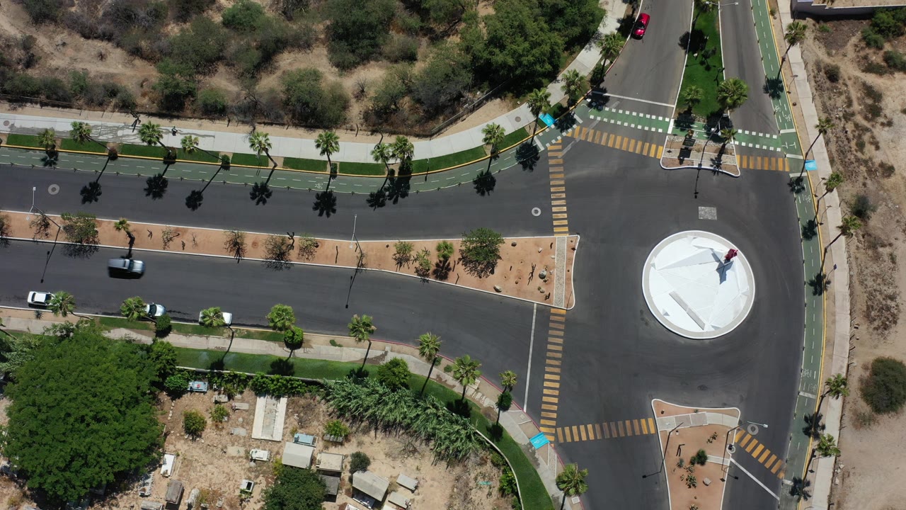Top-down aerial frame in Los Cabos, Mexico, showing a road junction, a white circular roundabout feature, crosswalks, bike lanes, palm shadows, cars, and no visible people.