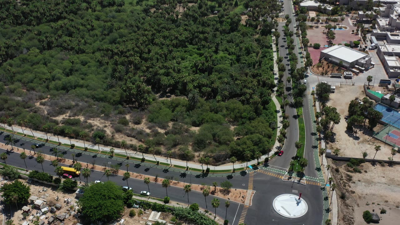 Aerial frame over Los Cabos, Mexico showing a roundabout, intersecting roads, dense green trees, a few cars, and no visible people.