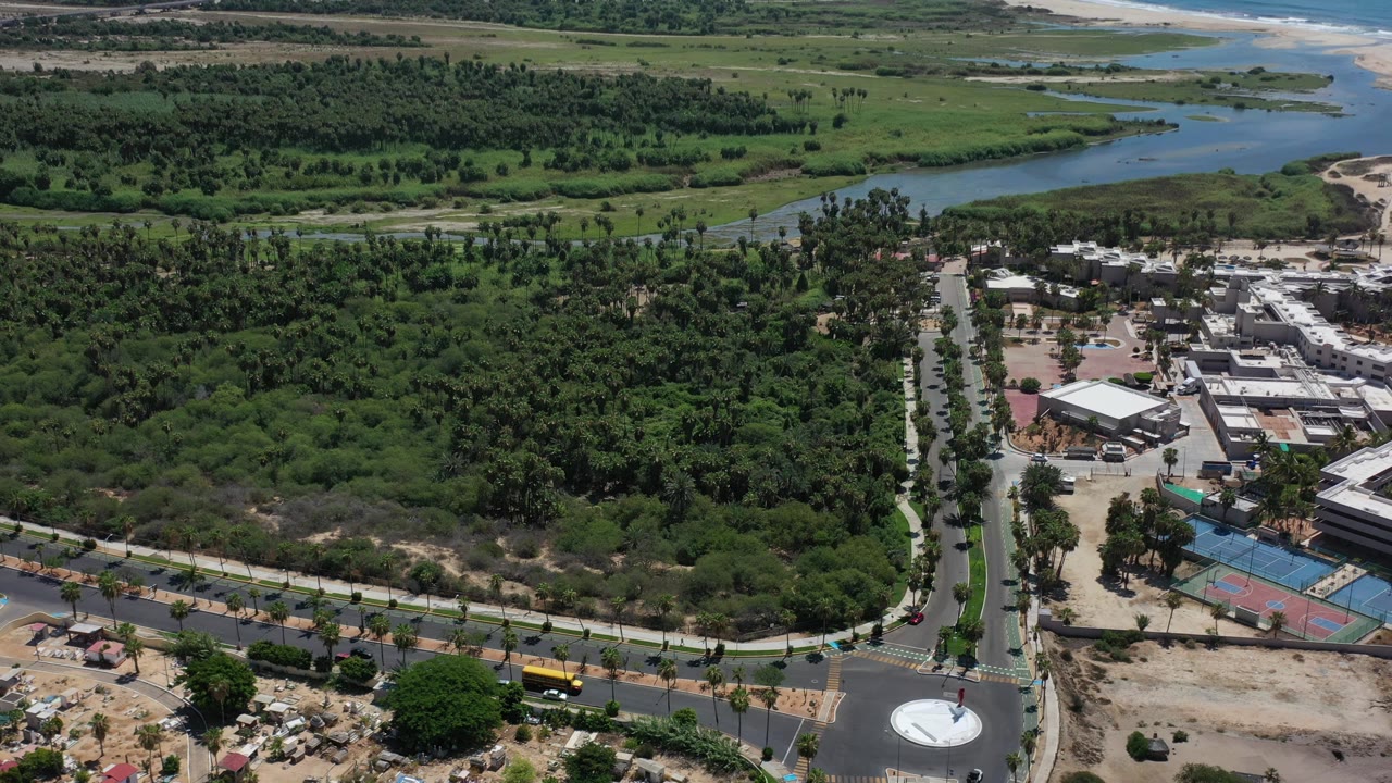 Aerial view across greenery toward a blue canal, sandy beach, ocean, and low buildings.
