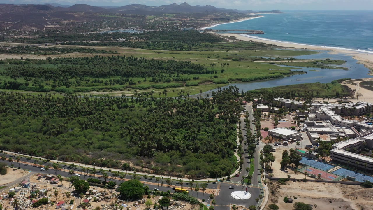 Aerial coastline with ocean, beach, lagoon, roads, buildings, palms, and hills.