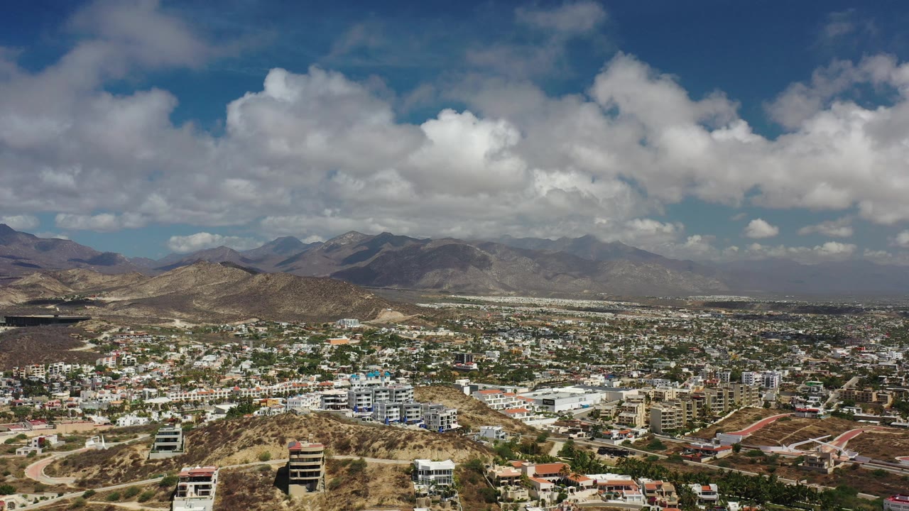 Wide aerial cityscape with dry hills, distant mountains, and cloudy sky.