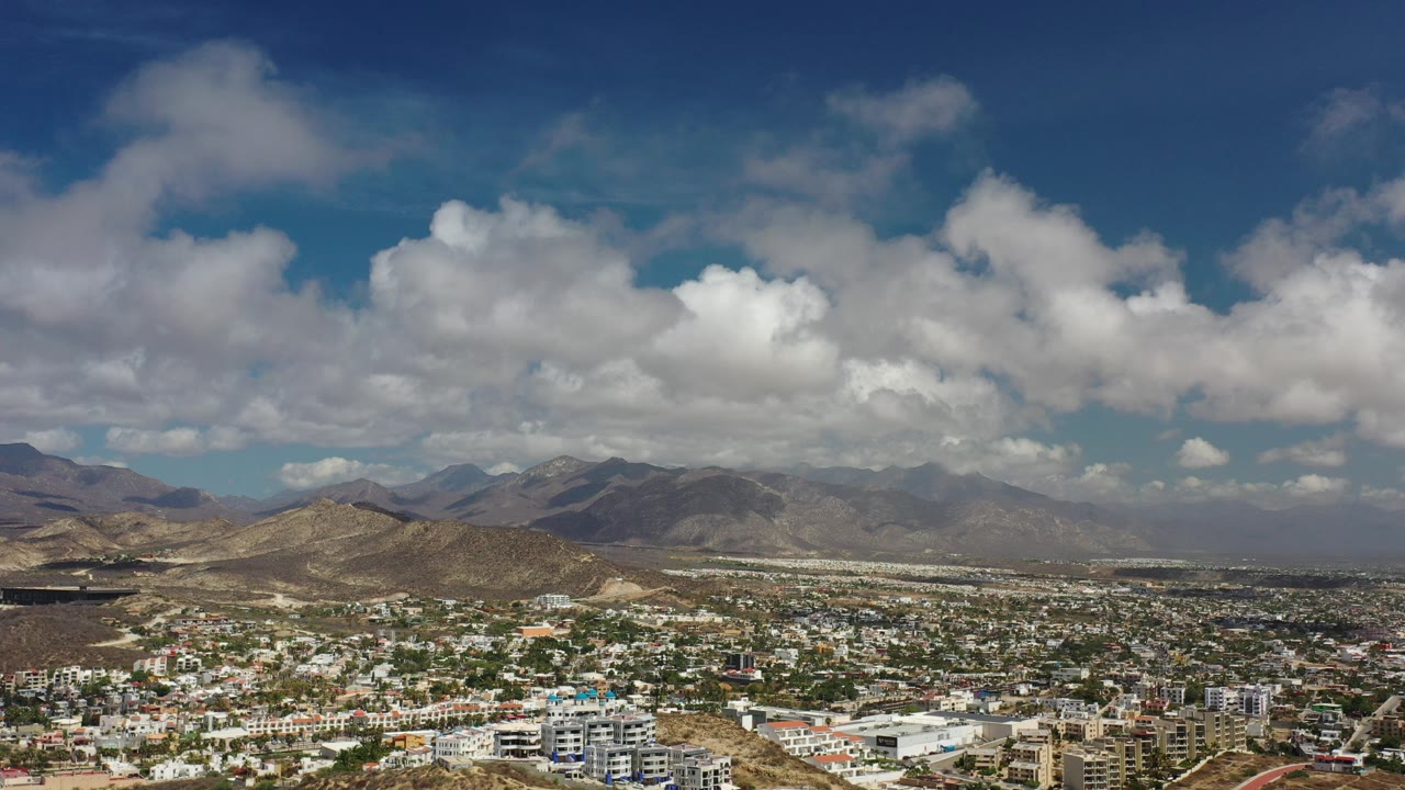 Opening frame of Los Cabos, Mexico from an elevated viewpoint, with a dense city at the bottom, dry mountain ridges in the distance, large clouds overhead, and no visible people.