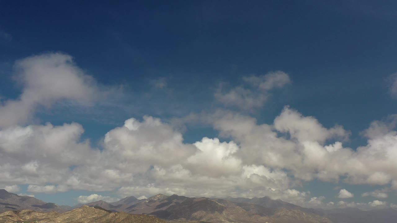 Los Cabos, Mexico frame with the mountain line still low in view and a bright central cloud mass spread across the blue sky, with no visible people.