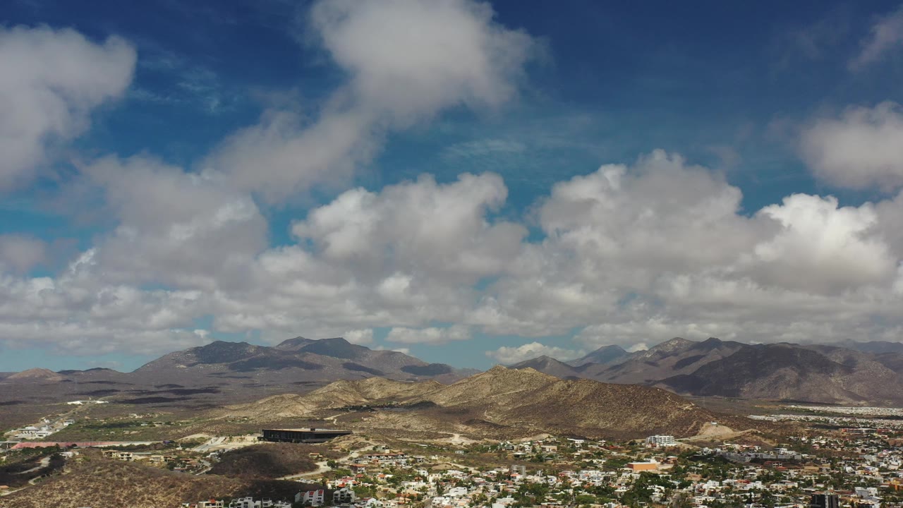 Opening frame of Los Cabos, Mexico, showing a broad blue sky with scattered white clouds above dry hills, distant mountains, and low town buildings, with no visible people.