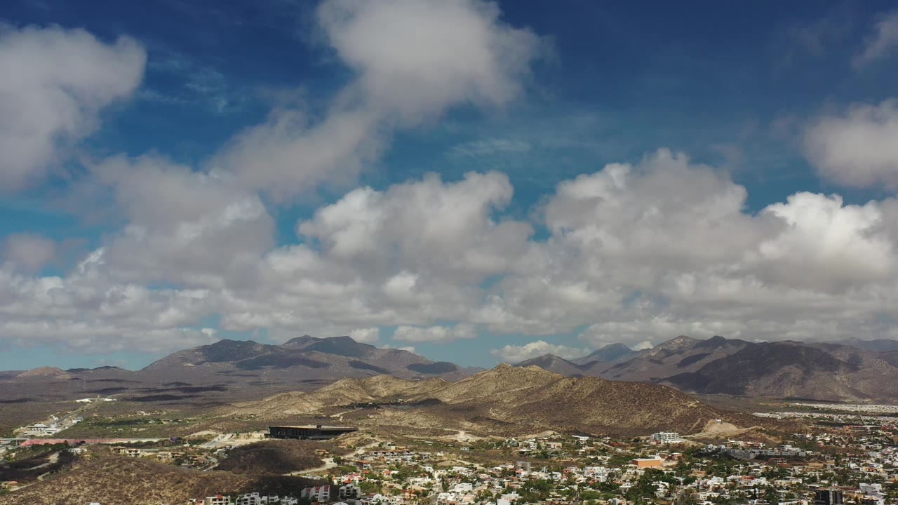 Bright clouds above mountains, dry terrain, and scattered buildings.