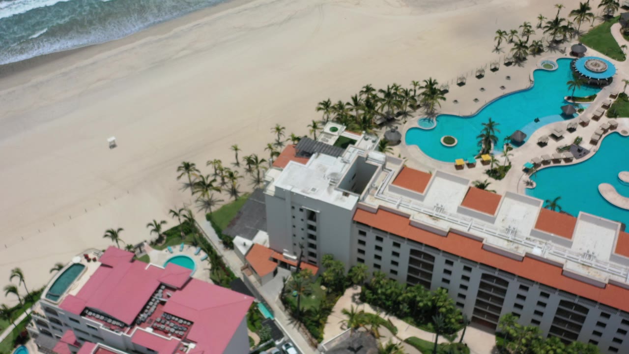 Aerial view of a beachfront hotel, pools, sand, and surf.