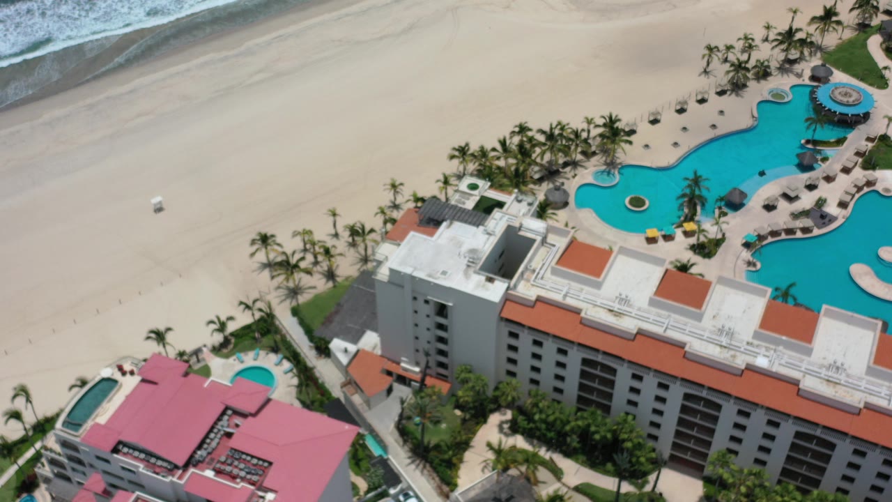 High aerial view over a Los Cabos resort with red and white rooftops, a curved blue pool, palm trees, pale beach, breaking waves, and no visible people.