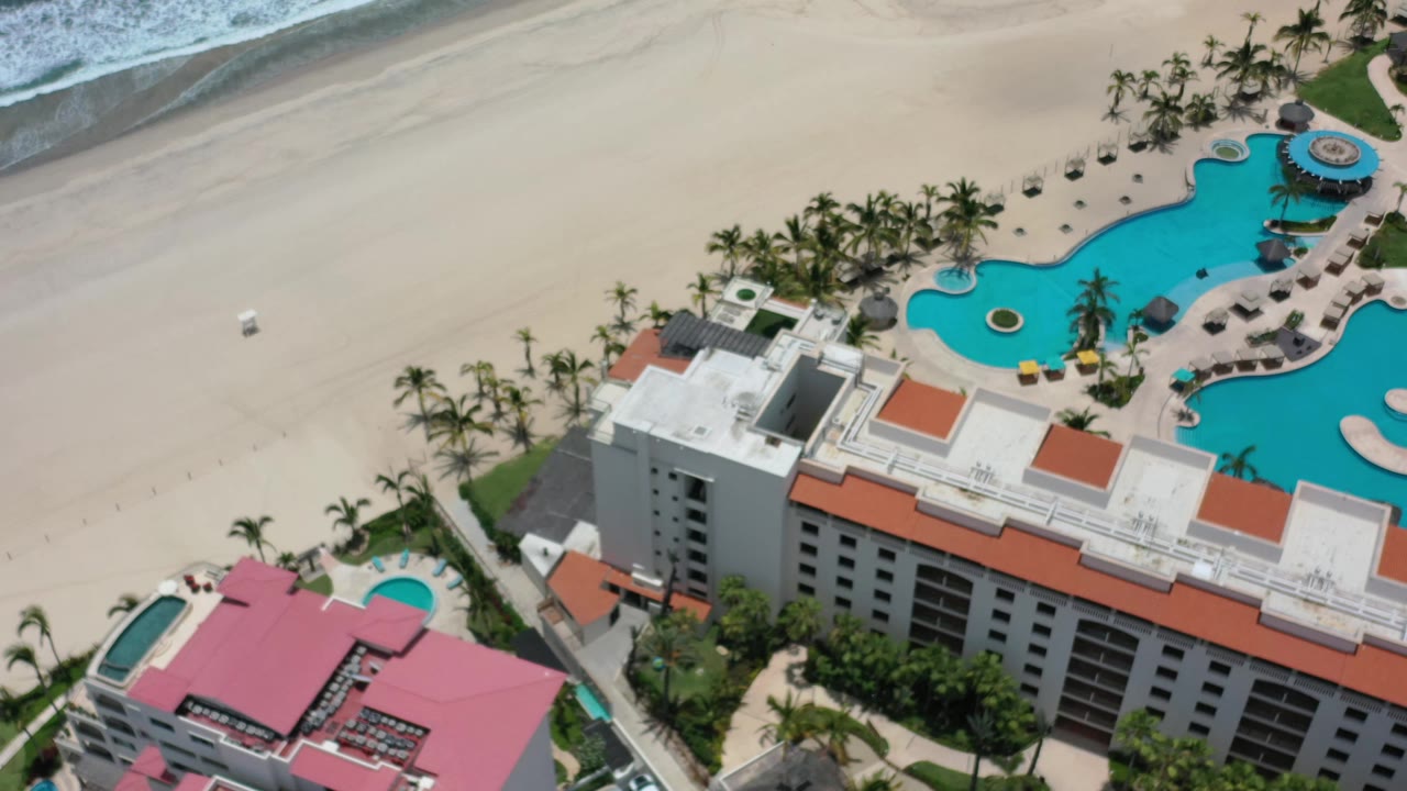 Aerial frame in Los Cabos showing the resort roofline lower in view, the turquoise pool to the right, open sand beyond it, surf at the top, and no visible people.