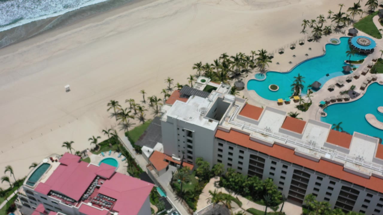 Drone-like view of beach resort, palm trees, pool, sand, and ocean edge.