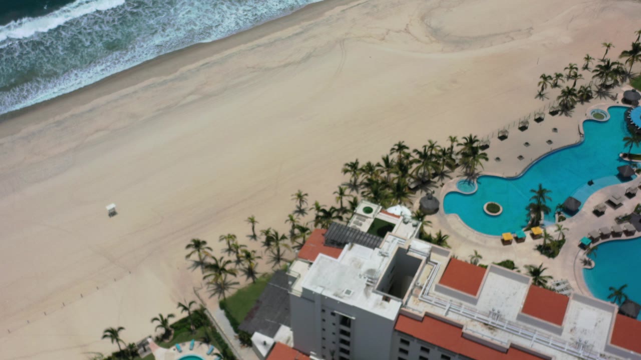 Overhead view of beach, resort pool, palms, and hotel roof.