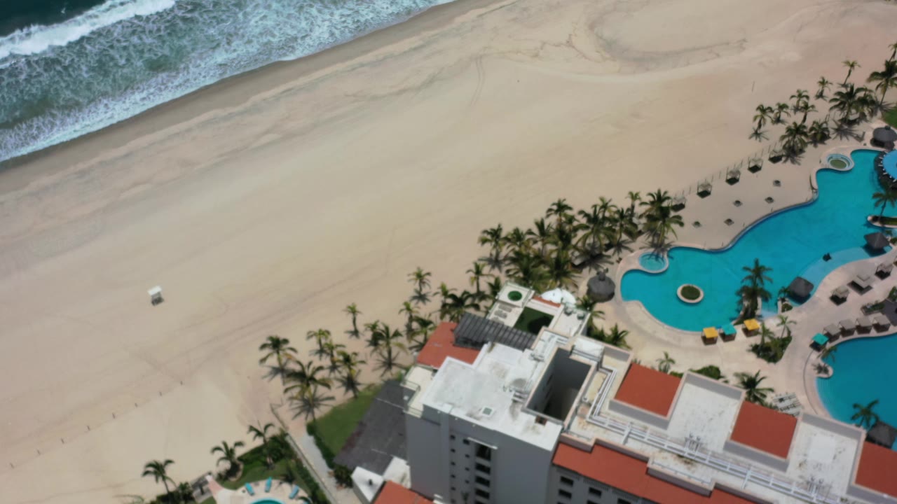 High-angle Los Cabos resort view with the sandy beach spanning the frame, waves at the top, palms around the pool, and no visible people.