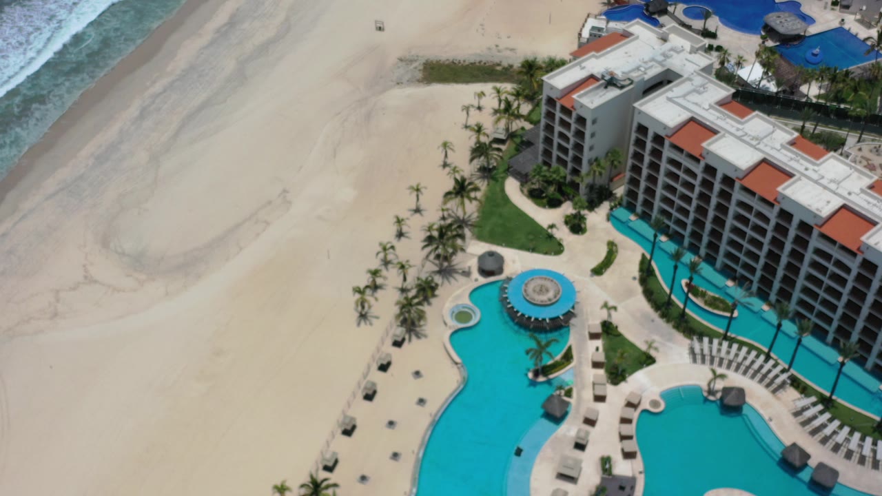 Aerial view of beach, surf, pools, palms, and beachfront buildings.