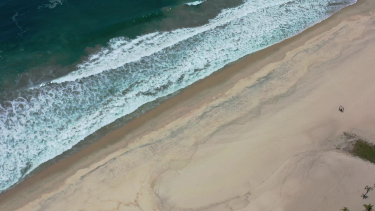 Overhead view in Los Cabos, Mexico, showing teal surf washing diagonally across pale sand with no visible people.