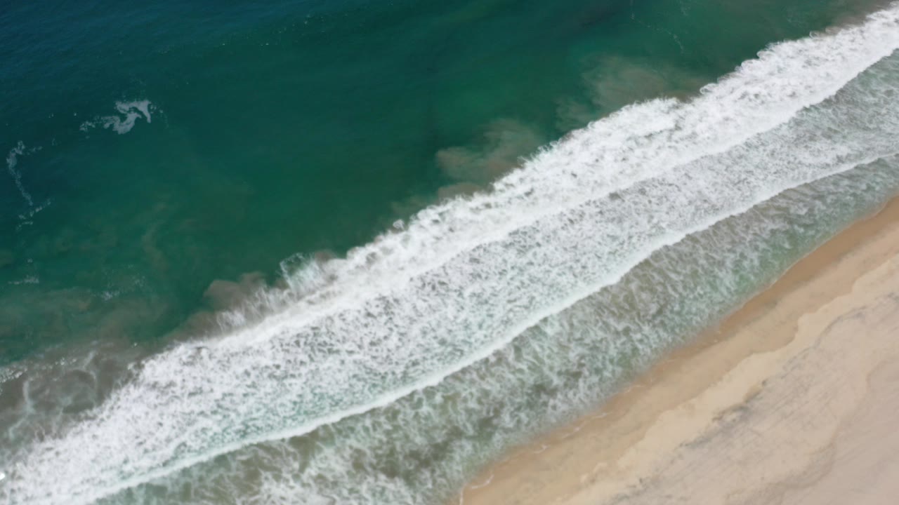 Aerial view of white surf reaching tan sand beside teal water.
