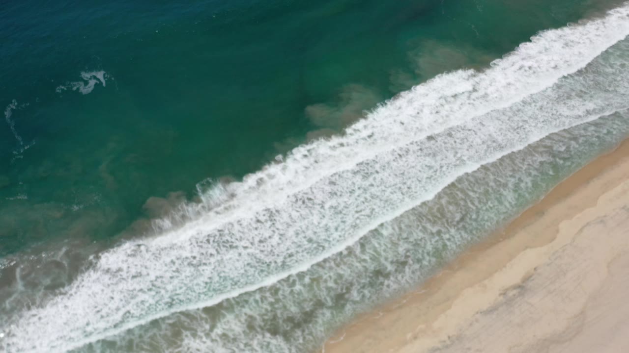 Aerial Los Cabos shoreline frame with foamy white water spreading over the shallows and tan beach remaining clear of visible people.