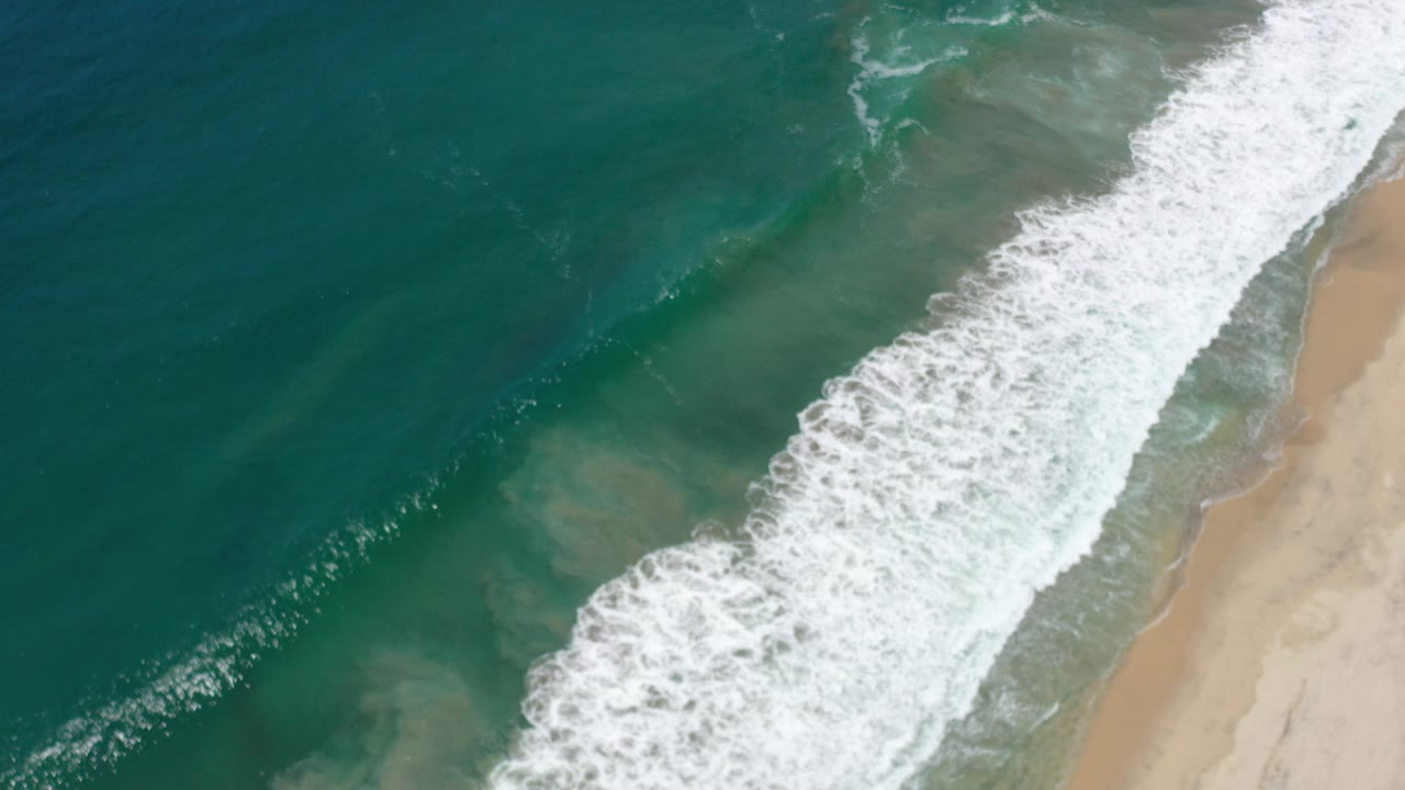 Aerial frame over Los Cabos, Mexico shows a broad white surf band breaking diagonally beside teal water and a strip of sand, with no visible people.