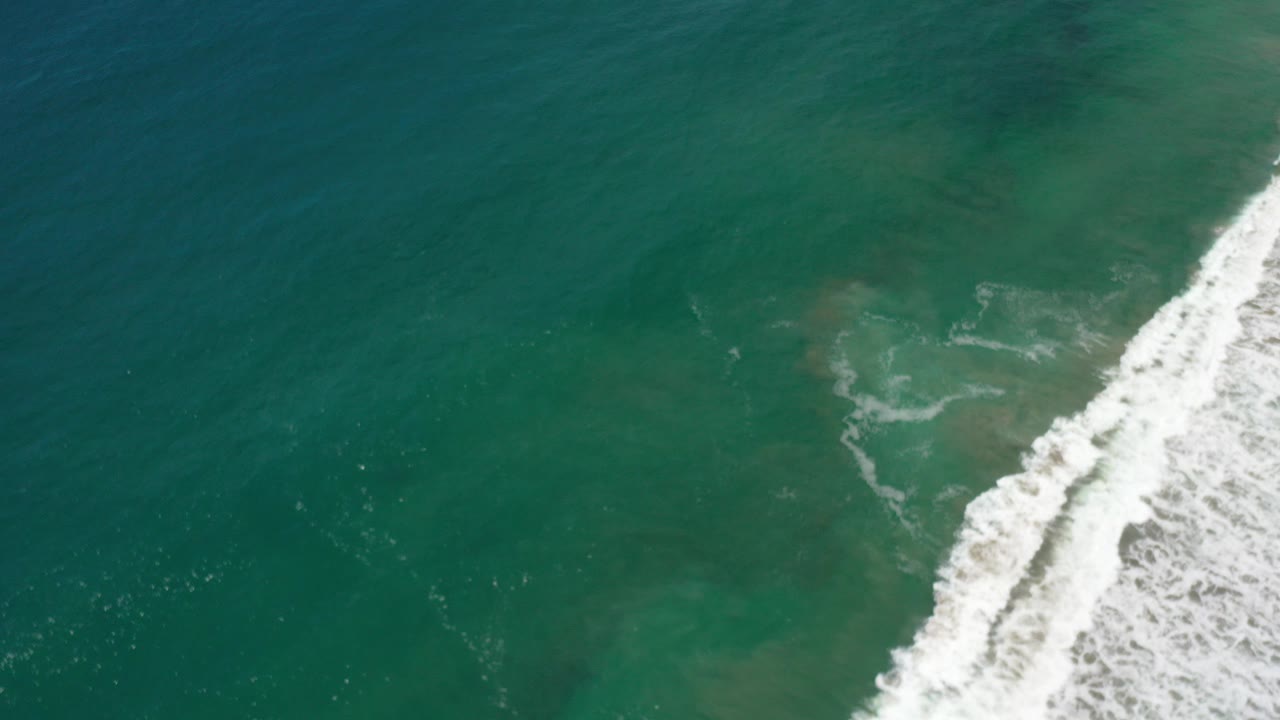 Overhead view of green-blue ocean in Los Cabos, Mexico, with white shoreline surf entering along the right edge and no visible people.