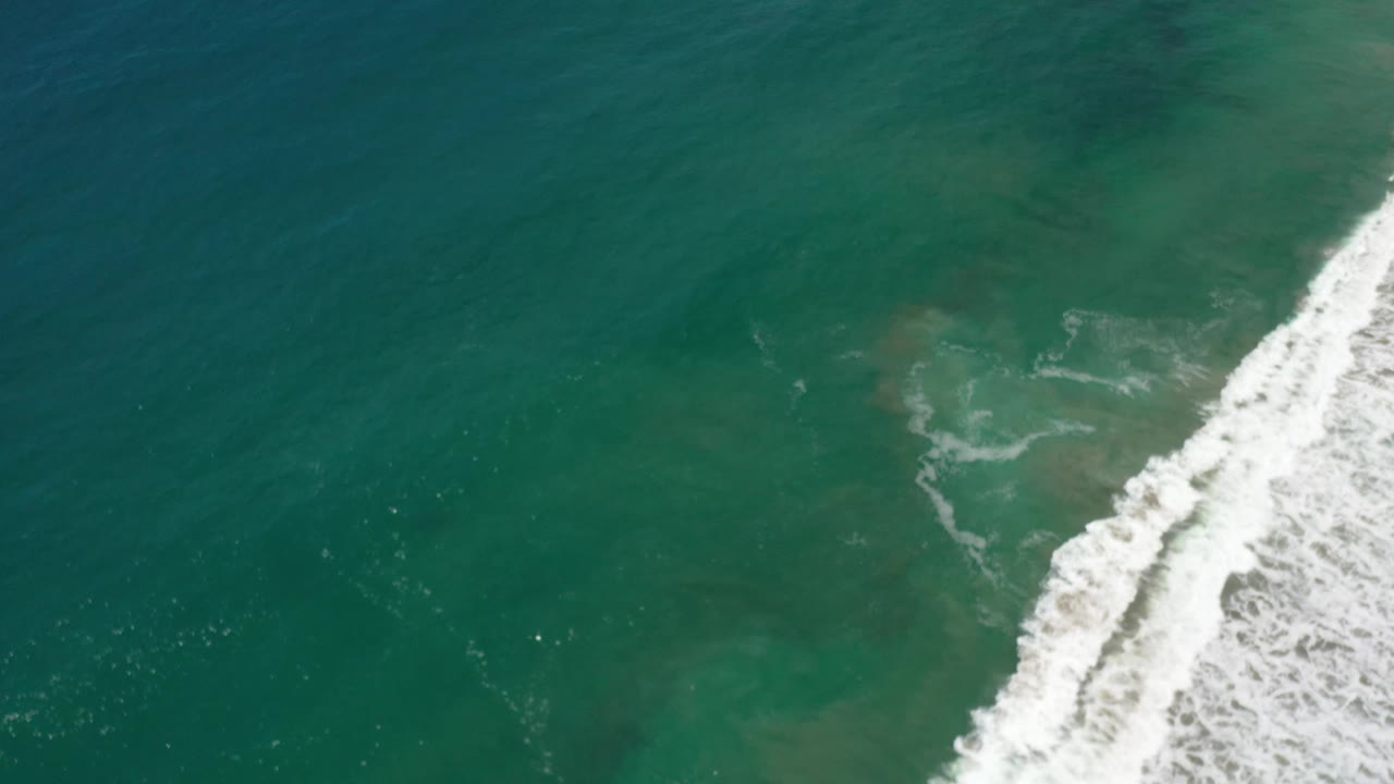 Aerial view over green ocean in Los Cabos, Mexico, with a diagonal band of white wave foam near the right side and no visible people.