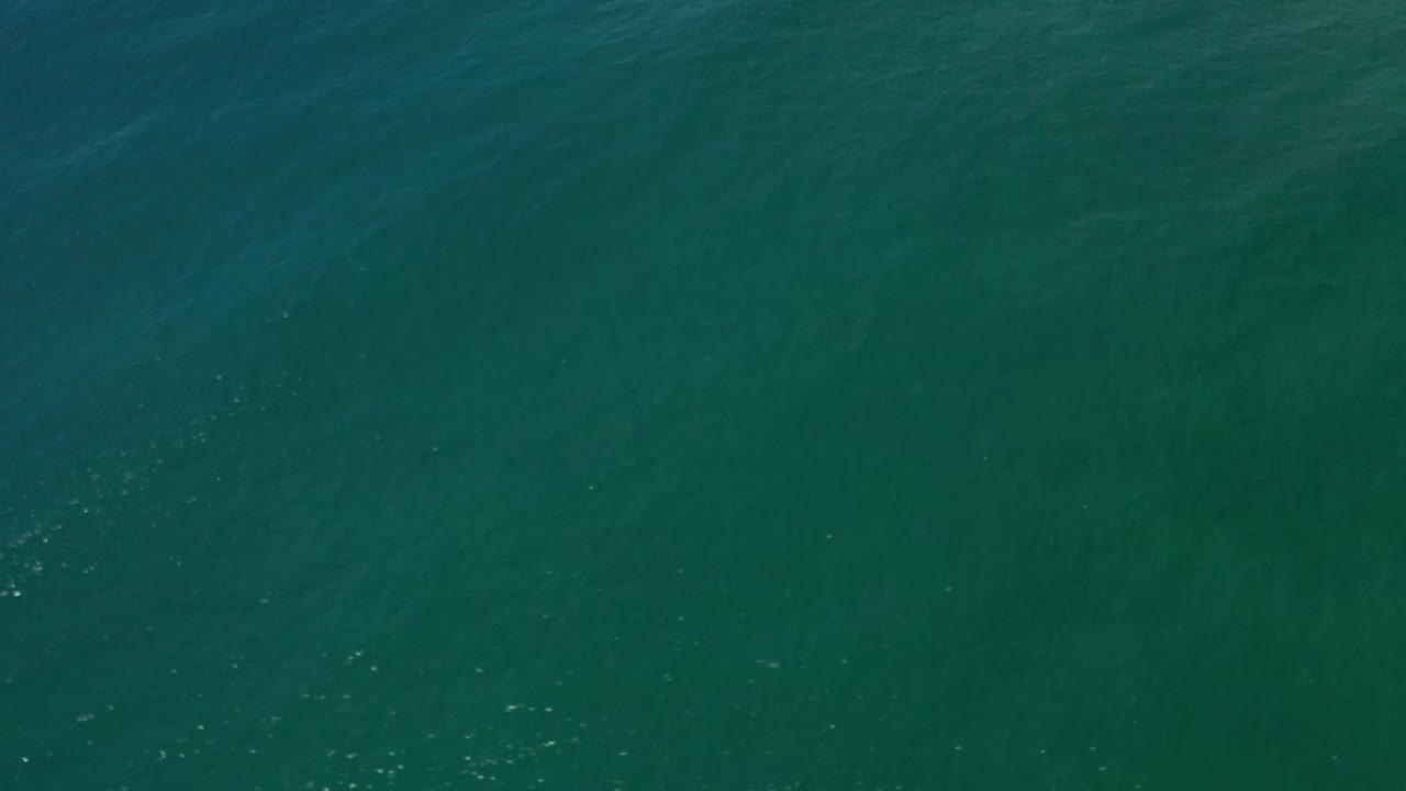 Overhead Los Cabos ocean frame showing blue-green water, muted surface bands, and a few bright specks from daylight on the ripples, no visible people.