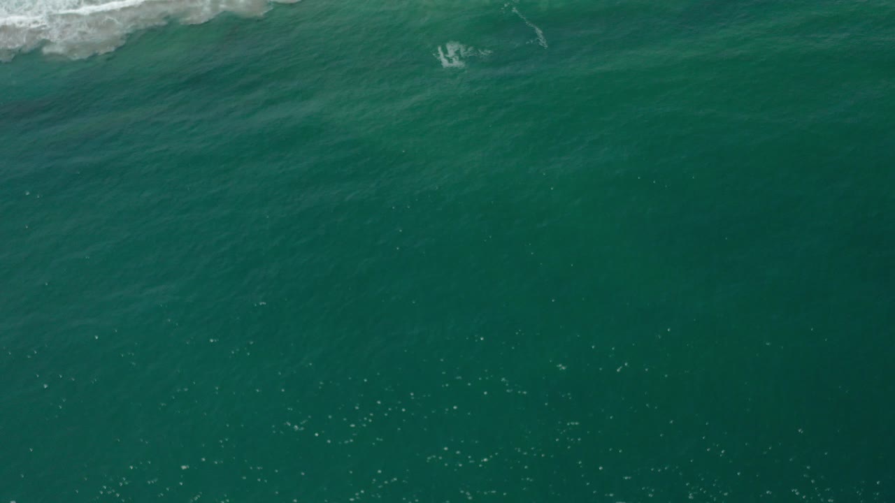 Aerial view over teal water in Los Cabos, Mexico, with thin white surf along the top edge and no visible people.