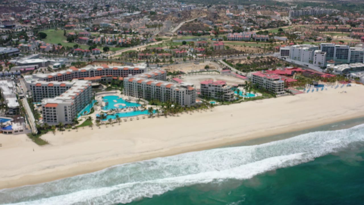 High aerial view over a Los Cabos beachfront resort, with white surf in the foreground, blue pools beside hotel buildings, town blocks behind, and no visible people.