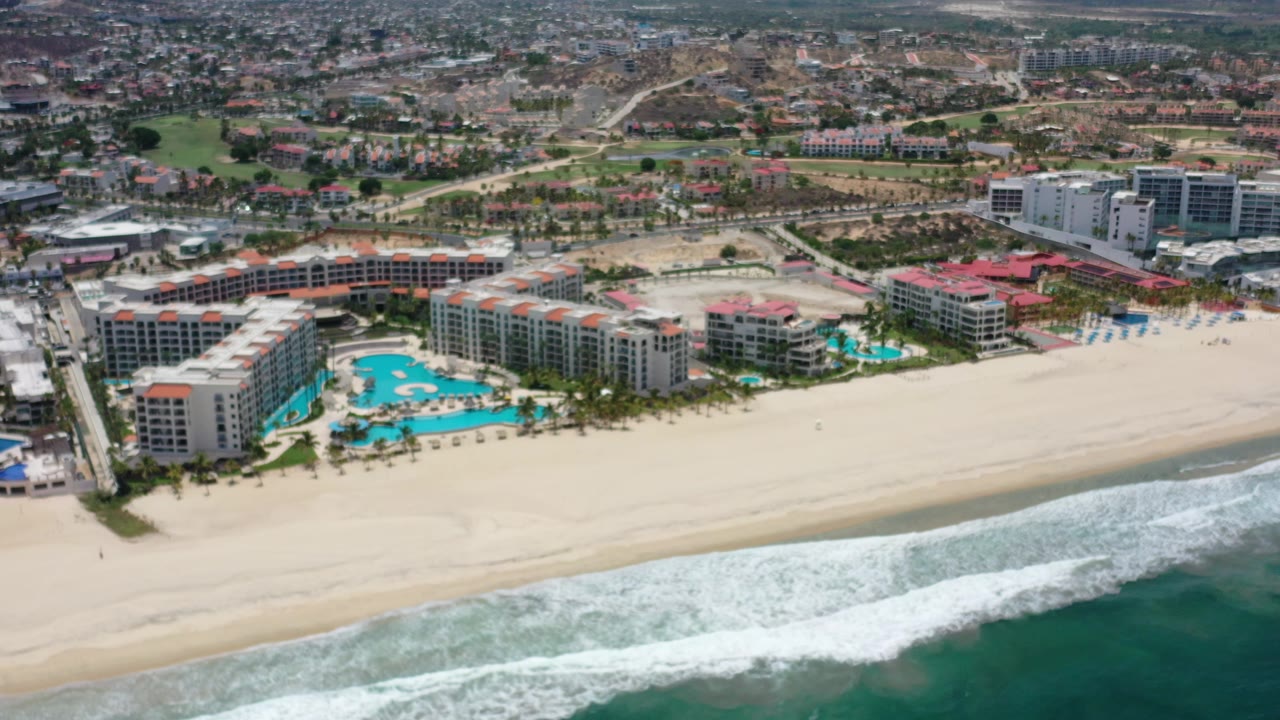 Wide aerial of resort buildings, beach, surf, town, and hills.