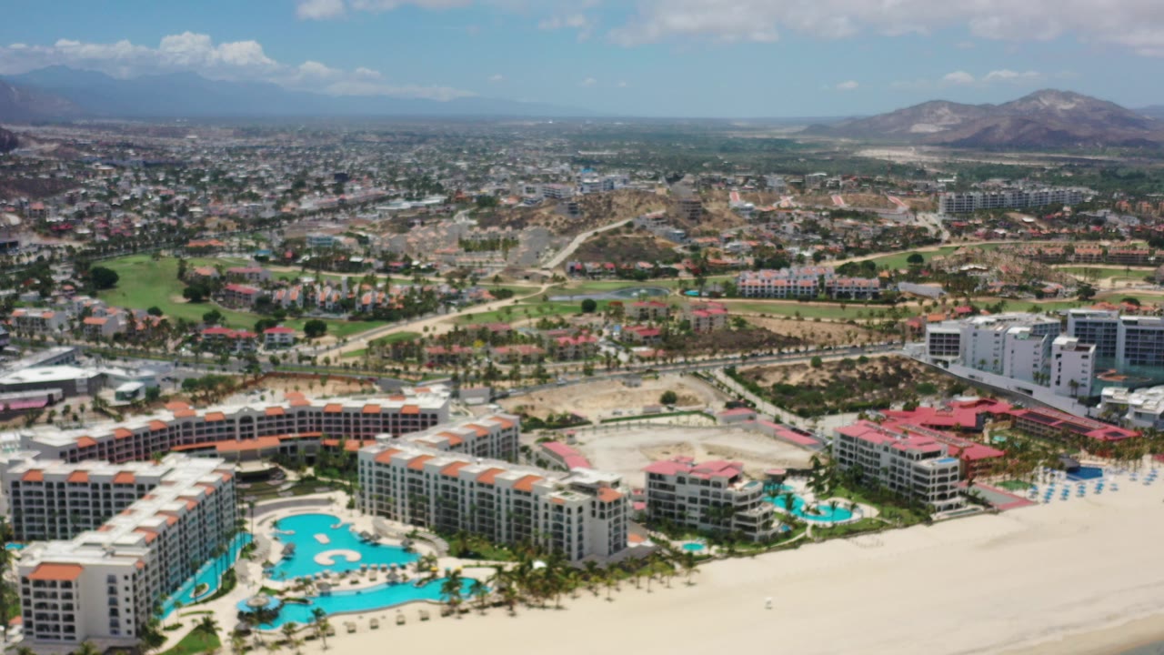 Aerial view of beachfront resort buildings, pools, sand, town, and hills.
