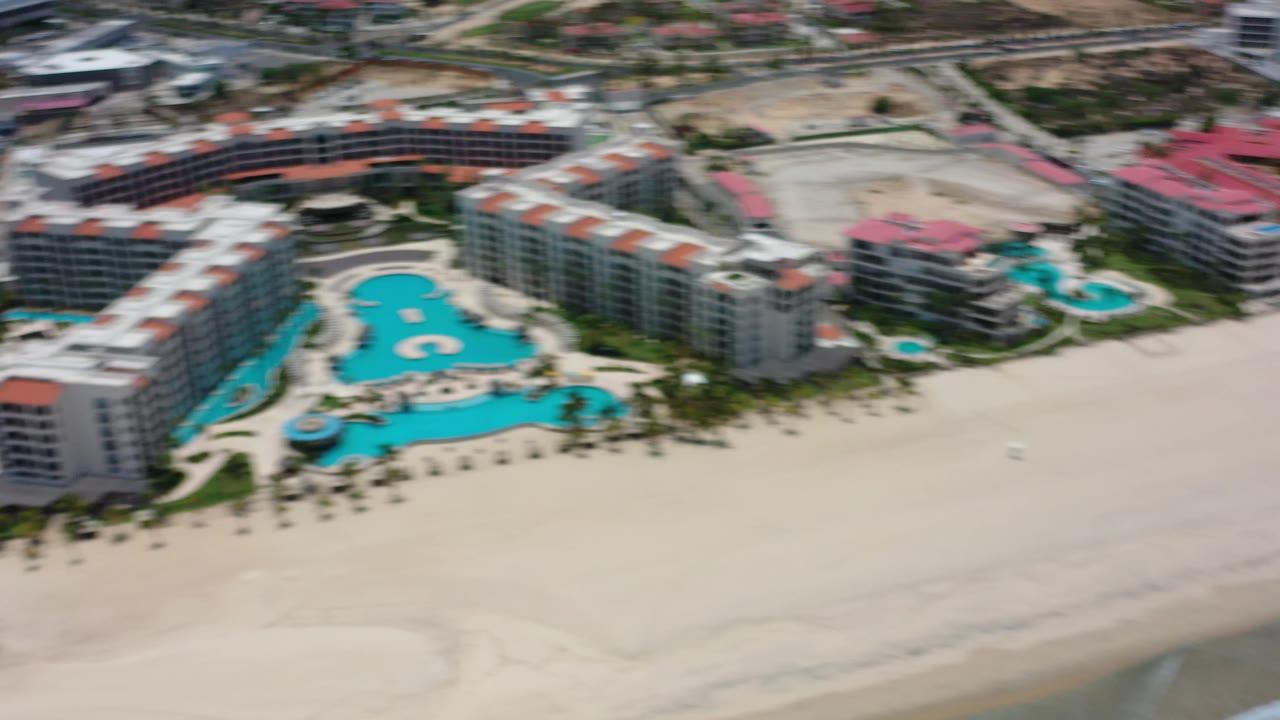 Aerial Los Cabos, Mexico frame with visible motion blur across the resort buildings, blue pool, sandy beach, and surf edge.